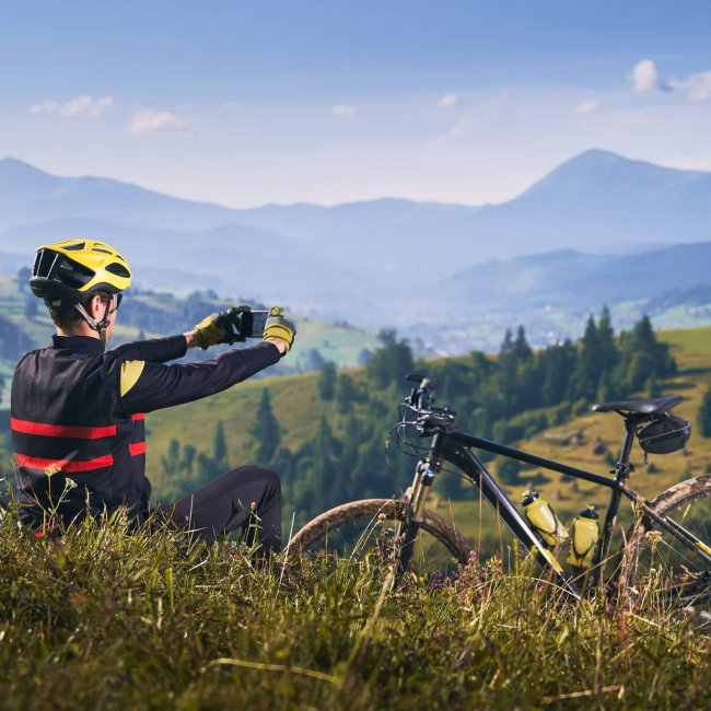 Young man sitting on grass near bicycle and talking mountain photo with modern smartphone. Male bicyclist enjoying the view of majestic mountains. Concept of sport and mountain biking.